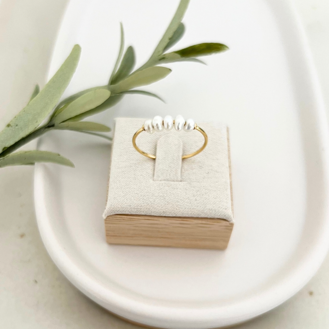 Goldener Ring mit Süßwasserperlen auf einem eleganten Präsentationskissen, dekoriert mit grünen Blättern, minimalistisch und zeitlos – ideal als hochwertiges Schmuckstück