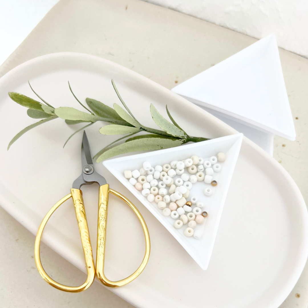 Verschiedene cremefarbene Perlen in einer Perlenschale, daneben eine goldene Schere – ideal für kreative DIY Projekte