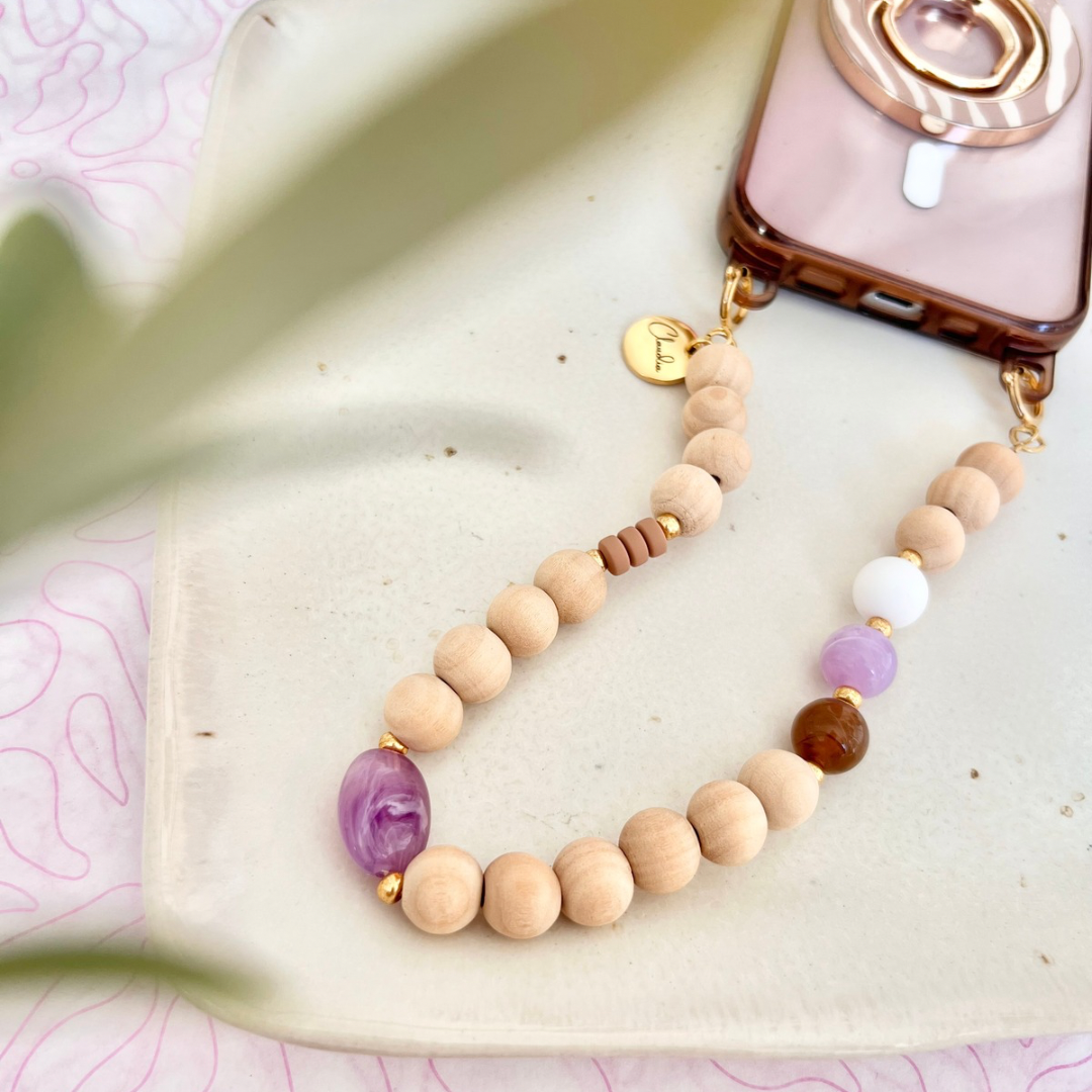Draufsicht auf eine im Workshop gefertigte Perlen-Handykette aus runden Holzperlen in Naturbeige, kombiniert mit einzelnen Glas- und Acrylperlen in Lila, Weiß und Braun sowie goldfarbenen Metallelementen und personalisiertem Edelstahl-Anhänger, befestigt an einem Smartphone auf hellem Untergrund mit Pflanzenblatt.