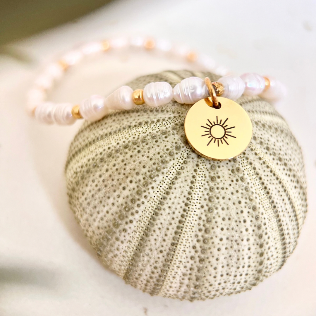 Ein elastisches Perlenarmband aus kleinen weißen Süßwasserperlen mit goldenen Zwischenelementen und rundem Sonnenanhänger liegt auf einer strukturierten Muschel