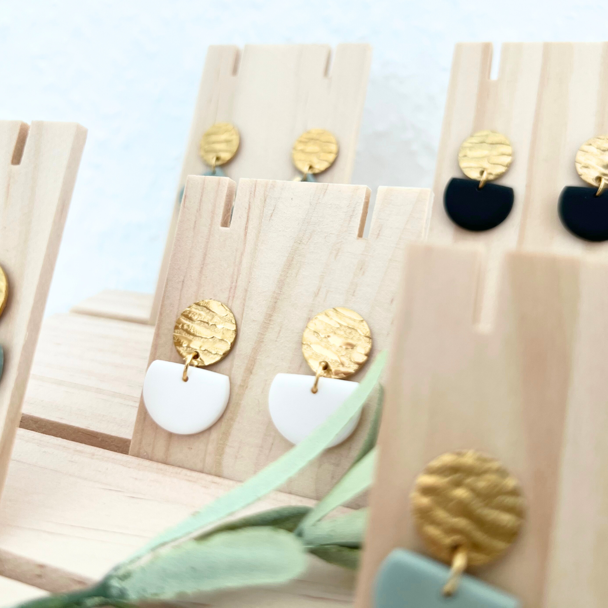 Mehrere Paar Ohrringe MURADA in verschiedenen Farben, mit Fokus auf die weißen Ohrringe aus Edelstahl und Polymer Clay, die elegante Halbkreis-Form zeigen