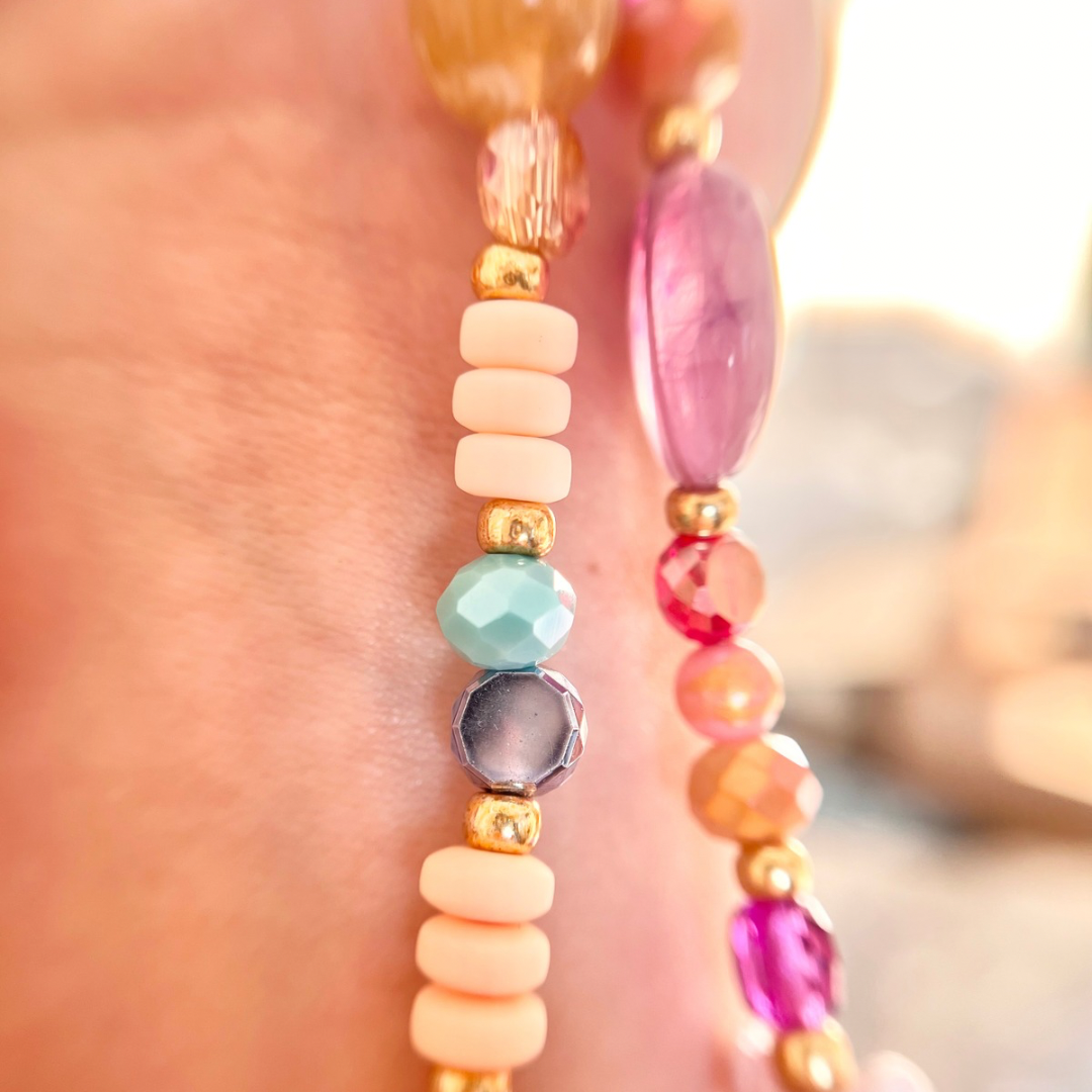 Makroaufnahme einer handgefertigten Perlen-Handykette mit ovalen und runden Perlen in Rosa, Weiß und Hellgrün sowie facettierten Glasperlen in Orange- und Rottönen, kombiniert mit kleinen goldfarbenen Metallelementen im warmen Licht.