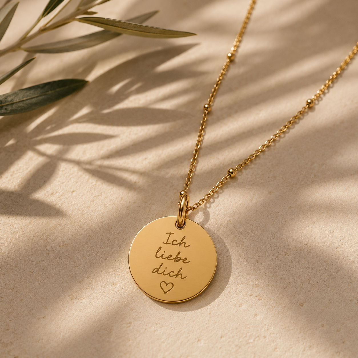 Nahaufnahme einer filigranen goldfarbenen Halskette mit rundem Gravur-Anhänger. Der Anhänger trägt die handschriftliche Gravur „Ich liebe dich“ mit Herzsymbol. Im Hintergrund sind dezente Schatten eines Olivenzweigs zu sehen.