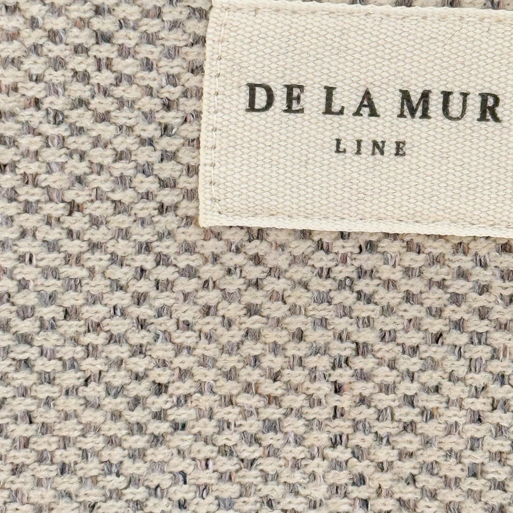 Nahaufnahme des beigen Möbelstoffs mit gewebter Struktur und DE LA MUR Label – handgefertigt in Barcelona.
