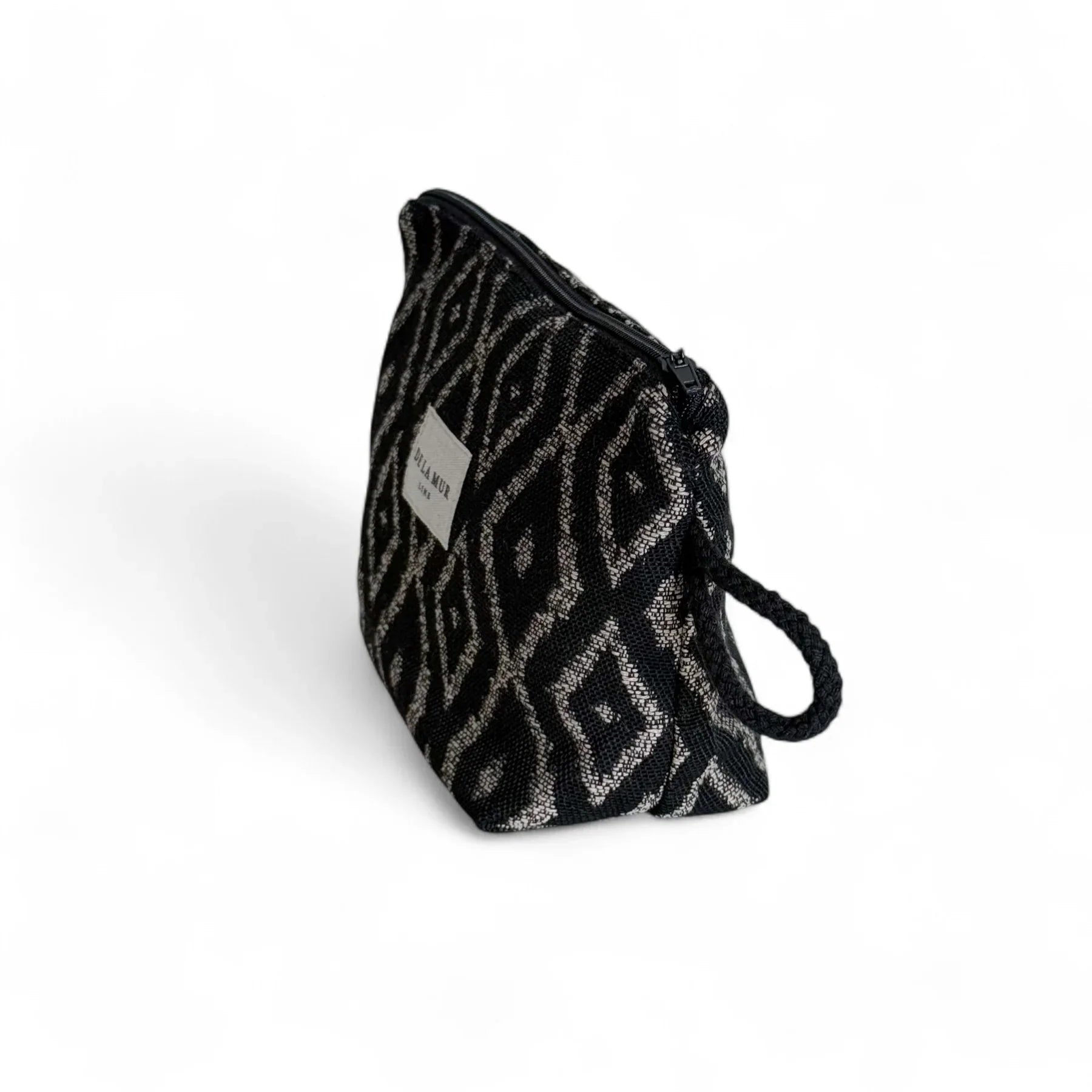 Eine geöffnete Kosmetiktasche mit schwarzem Jacquardstoff und beigefarbenem Diamantmuster liegt vor weißem Hintergrund. Das Innere ist dunkel gefüttert, der Reißverschluss geöffnet, seitlich befindet sich eine kleine Schlaufe.