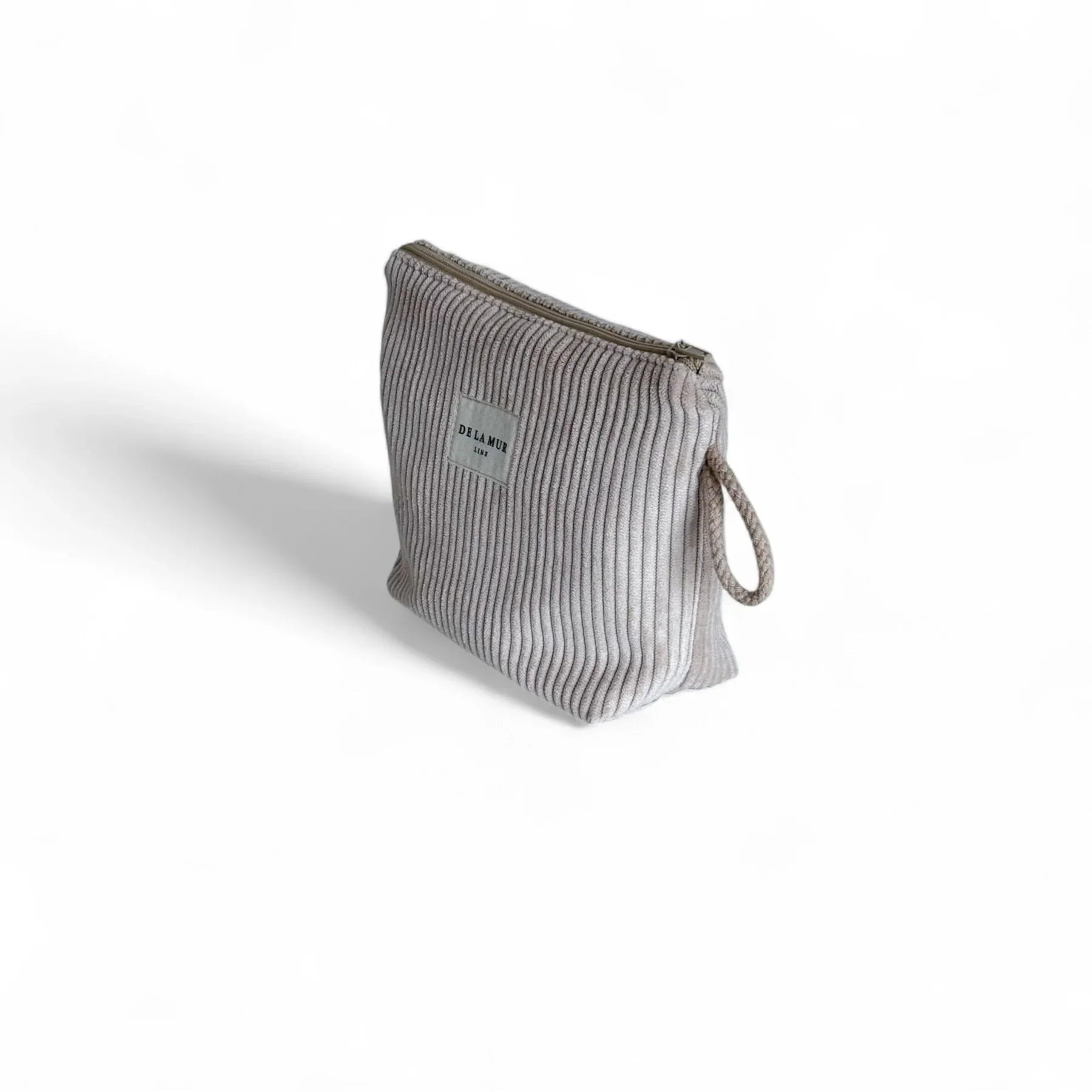 Eine kleine, taupefarbene Cordtasche steht im Dreiviertelwinkel auf weißem Hintergrund. Ein grauer Reißverschluss verläuft oben, seitlich hängt eine kurze geflochtene Schlaufe, vorne ist ein weißes DE LA MUR Label zu sehen. Idealer Organizer.
