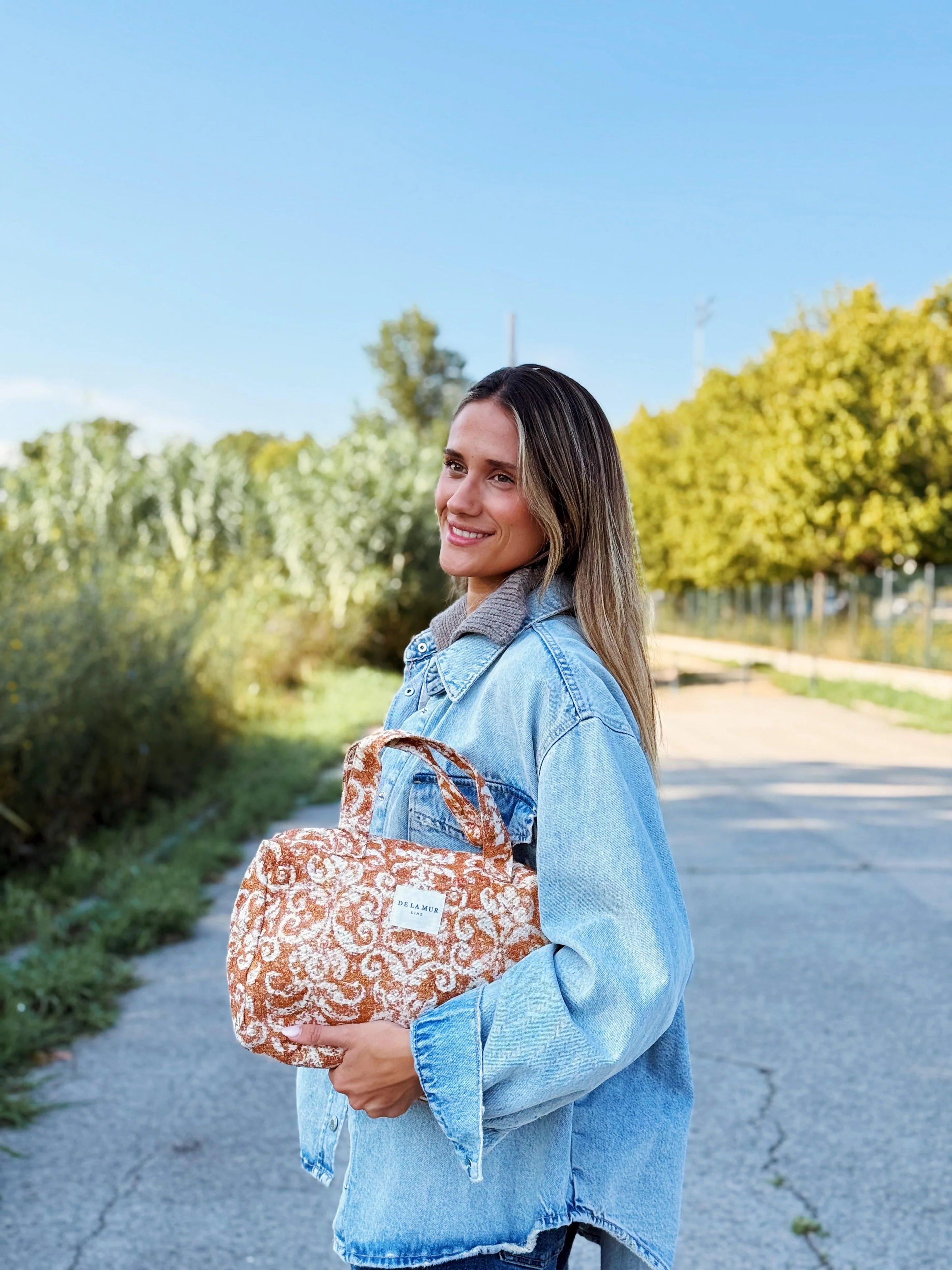 Eine Frau hält eine große Kulturtasche aus rostbraunem Jacquard-Stoff mit Ornamentmuster in der Hand. Die Beauty Bag hat zwei Tragegriffe, ein Innenfutter und ein Label mit der Aufschrift „DE LA MUR LINE“.