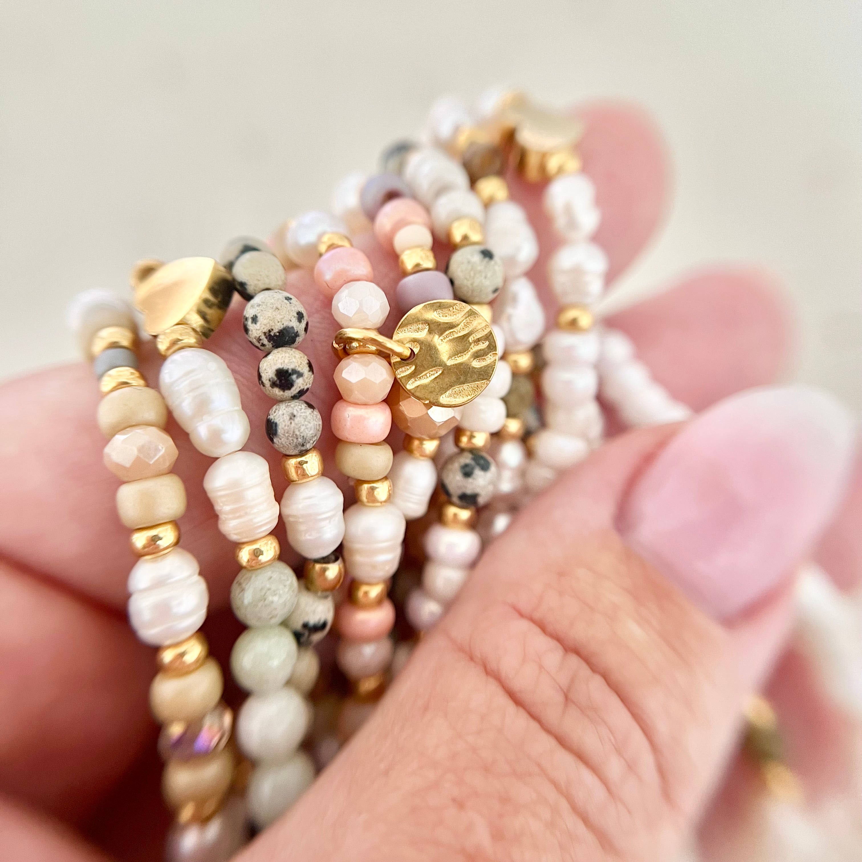 Hand hält mehrere Perlenarmbänder in sanften Naturtönen – Kombination aus Süßwasser-, Glas- und Natursteinperlen mit goldenen Anhängern.