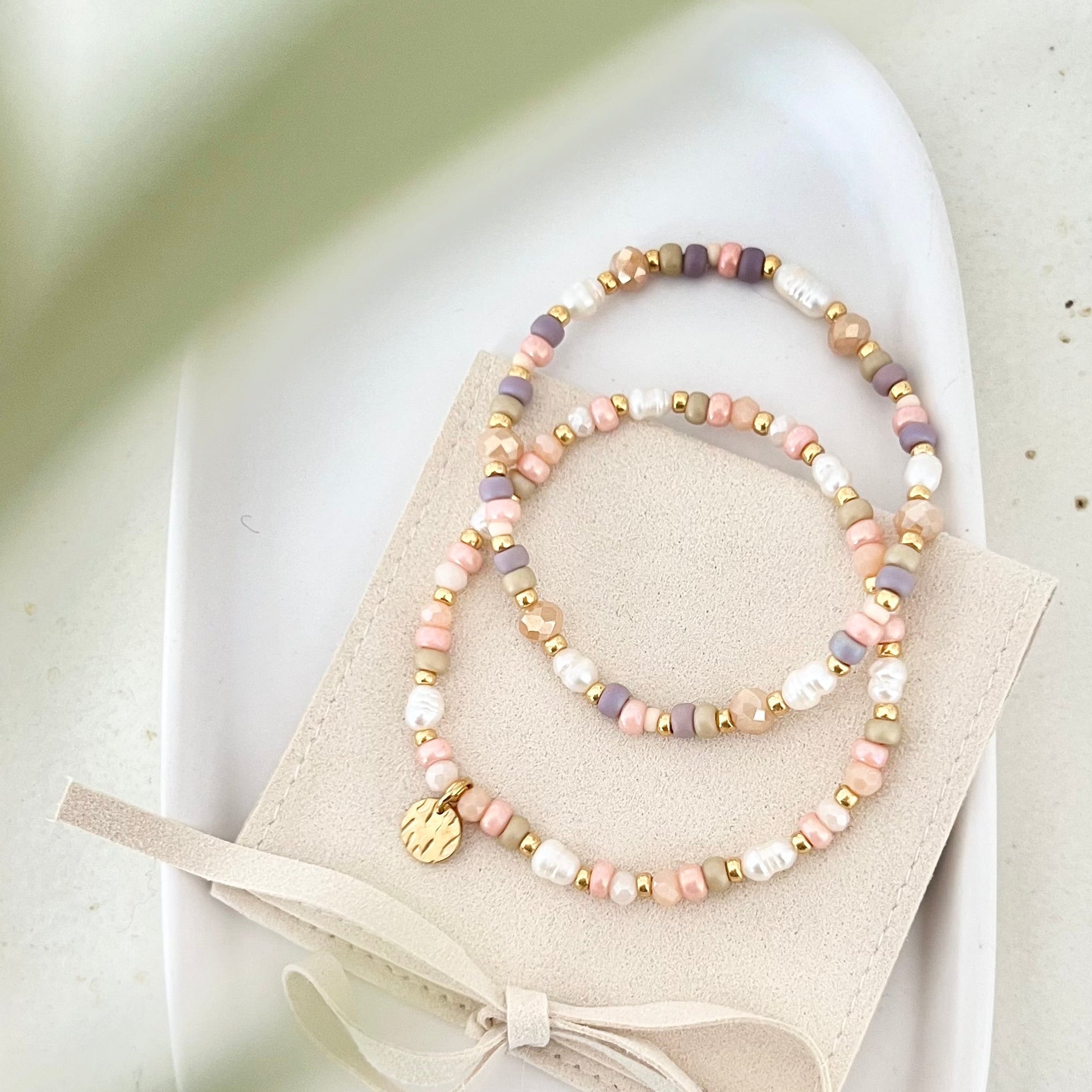 Zwei Armbänder auf einem beigefarbenen Schmuckbeutel, Pastellperlen und goldene Anhänger sichtbar, dekorativ auf Keramikschale platziert.