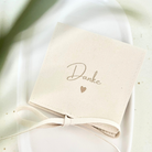 Beiger Schmuckbeutel mit der Aufschrift „Danke“ und Herzmotiv, edle Geschenkverpackung für Armbänder, Ketten oder Ohrringe. Nahaufnahme eines beigen Schmuckbeutels aus Mikrofaser mit Schleife und der Aufschrift „Danke“ mit Herz, liegend auf weißer Keramikschale