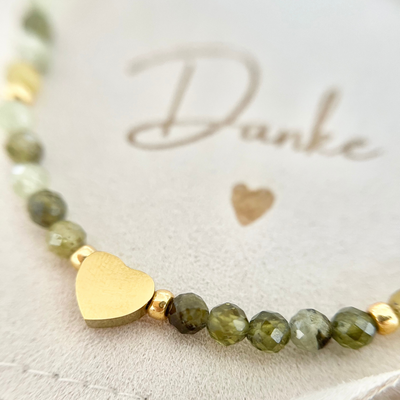 Nahaufnahme eines handgefertigten Perlenarmbands aus grünen Natursteinperlen mit vergoldetem Herz-Anhänger. Das Armband liegt auf einem Schmuckbeutel mit der Aufschrift „Danke“.