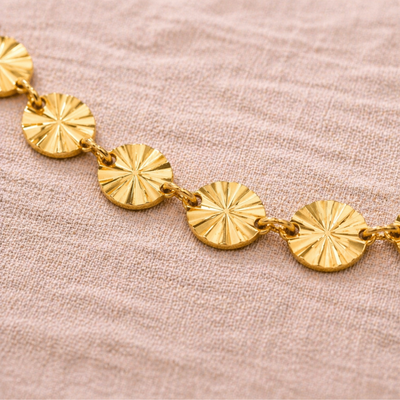 Detailaufnahme eines goldenen Edelstahl Armbands mit kleinen runden Sonnen-Elementen, glänzende Oberfläche auf beigem Stoff, hochwertiger wasserfester Schmuck