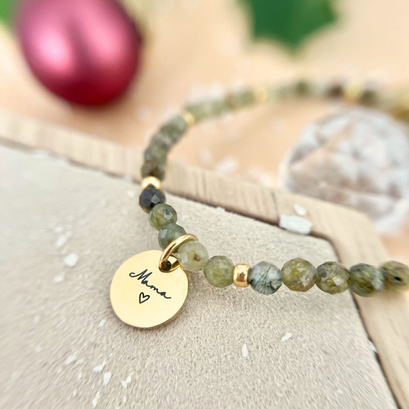 Detailaufnahme des Armbands auf heller Unterlage, goldener Gravur-Anhänger „Mama“ im Fokus, rote Weihnachtskugel im Hintergrund.