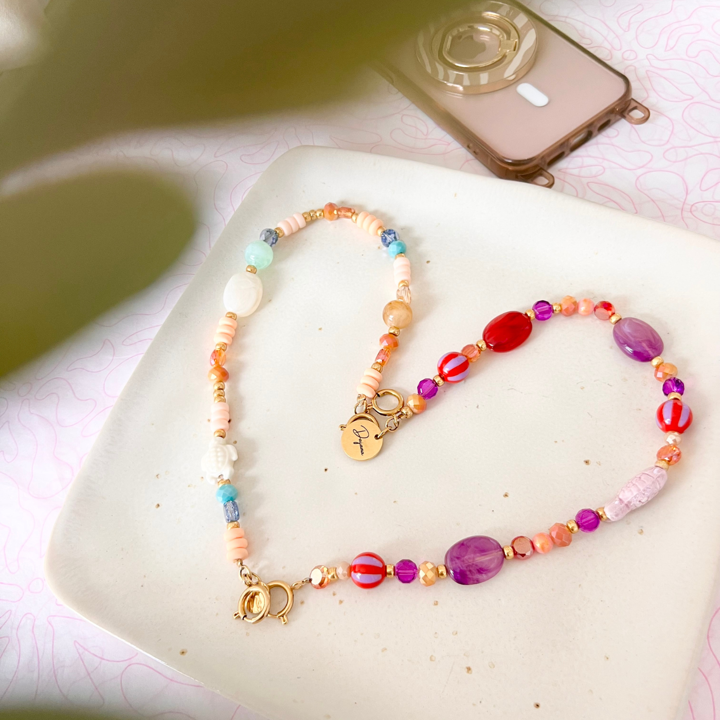 Gesamtansicht der Handykette LAGUNA mit Keramik-Schildkröte, bunten Glasperlen und goldenen Details neben einem Smartphone.