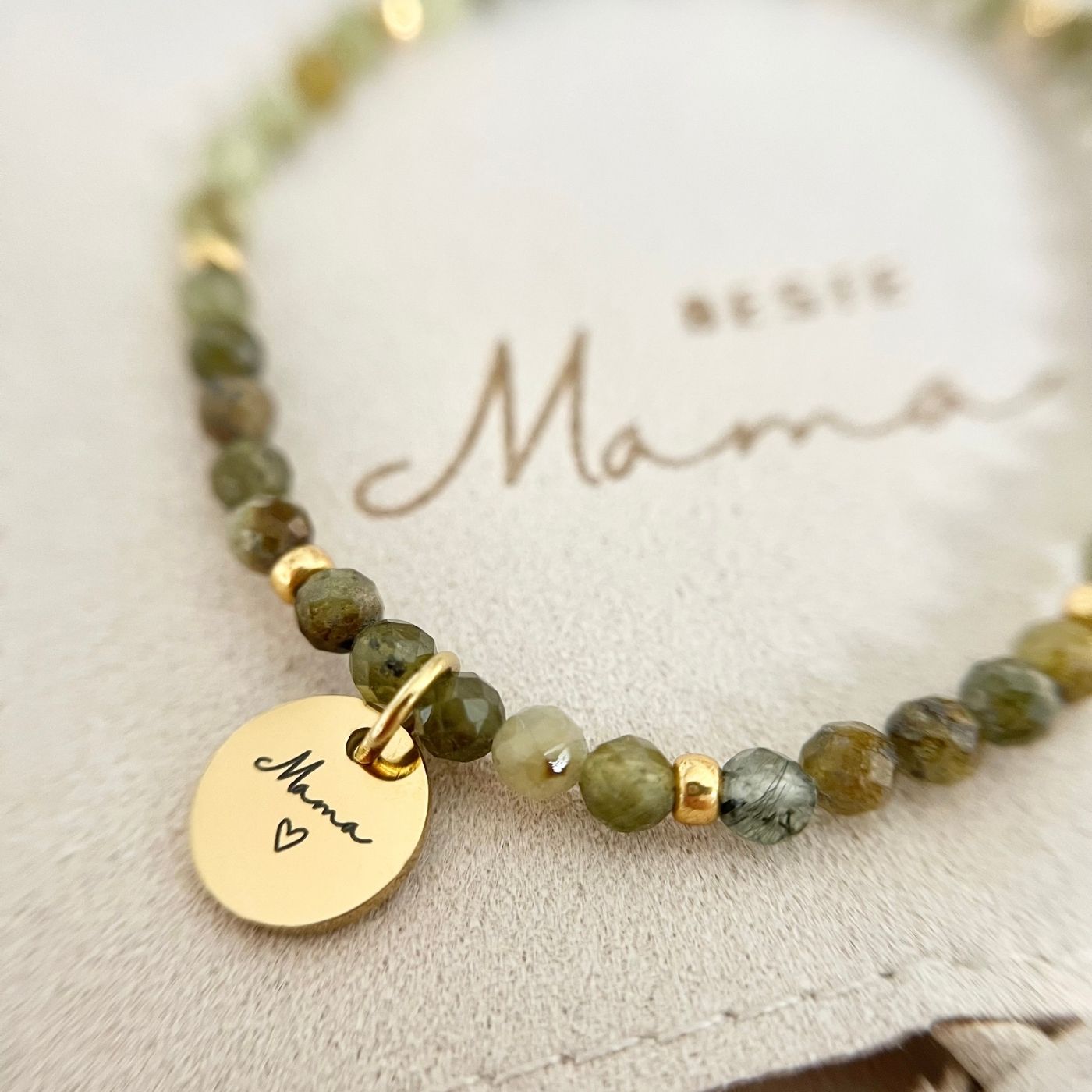 Nahaufnahme des Gravur-Anhängers „Mama“ mit Herz auf dem Perlenarmband, Hintergrund Schmuckbeutel „Beste Mama“.