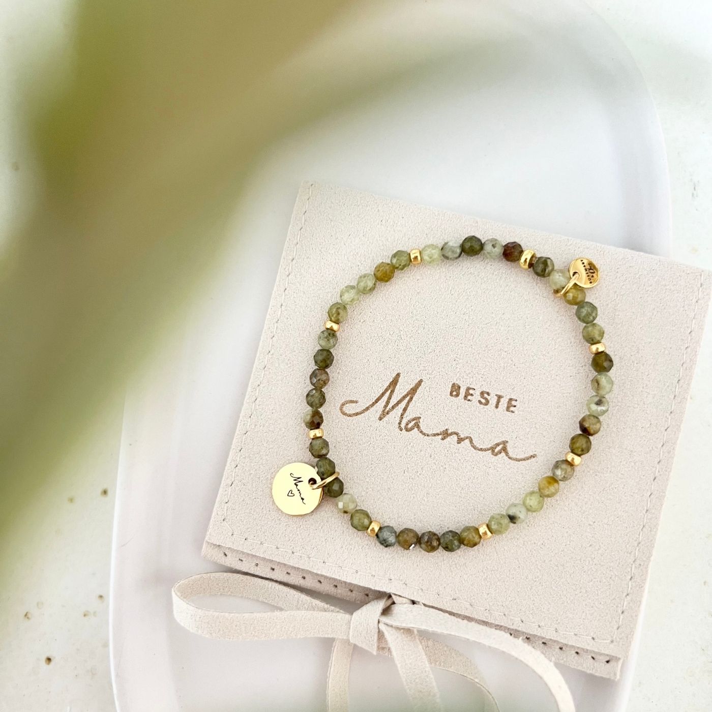 Armband mit Natursteinperlen in Grün und goldenen Akzenten, auf einem Schmuckbeutel mit Aufschrift „Beste Mama“ und rundem Gravur-Anhänger „Mama“ mit Herz.