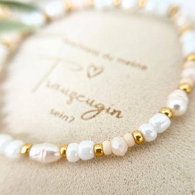 Detailansicht von Trauzeugin Schmuck – filigranes Armband mit glänzenden Perlen in Weiß, Nude und Gold, personalisierter Gravuranhänger mit Herzsymbol, auf beigem Schmuckbeutel mit gestickter Aufschrift „Trauzeugin“
