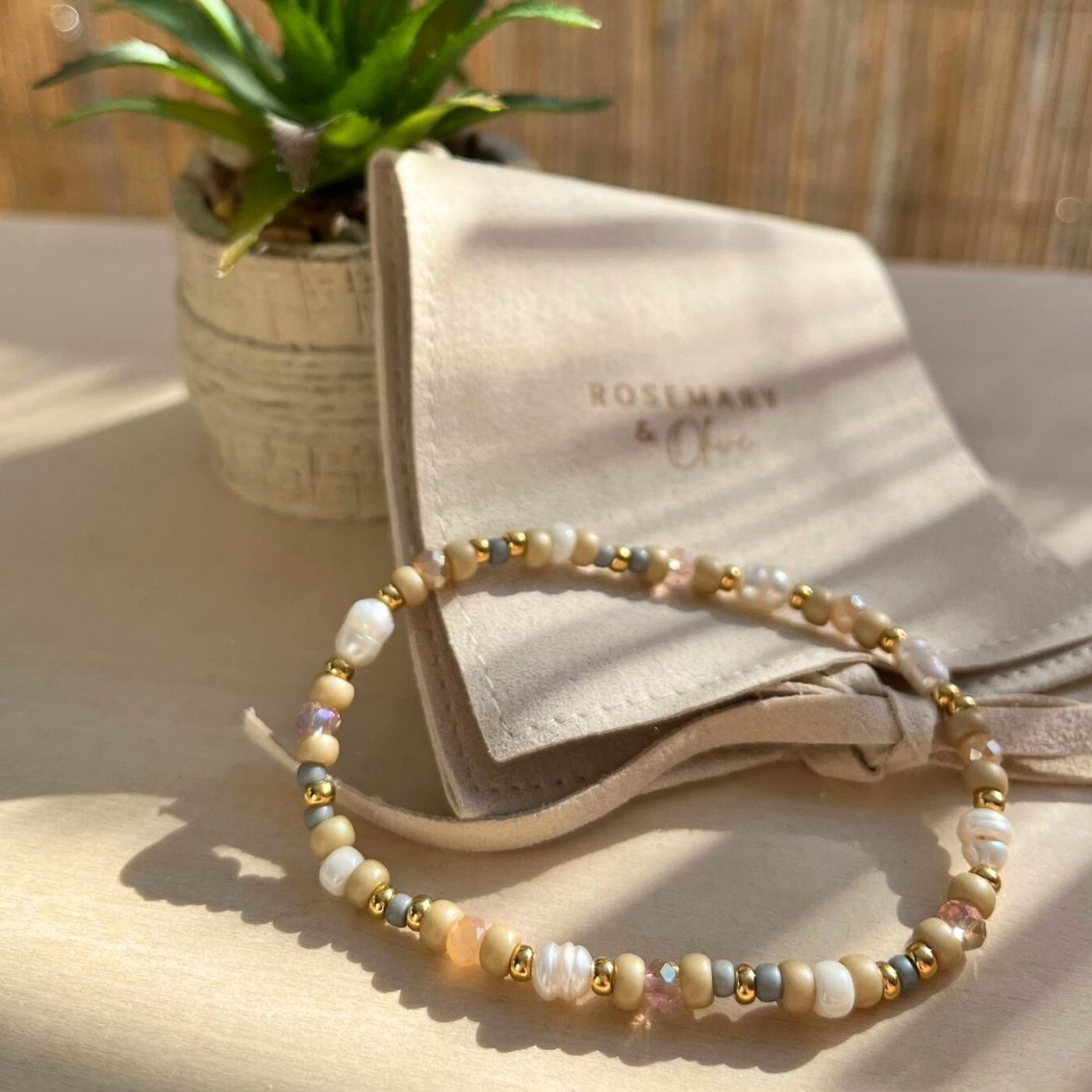 Boho Armband im Sonnenlicht auf heller Holzoberfläche, im Hintergrund ein Schmuckbeutel mit Logo „Rosemary & Olive“ und eine kleine Pflanze.