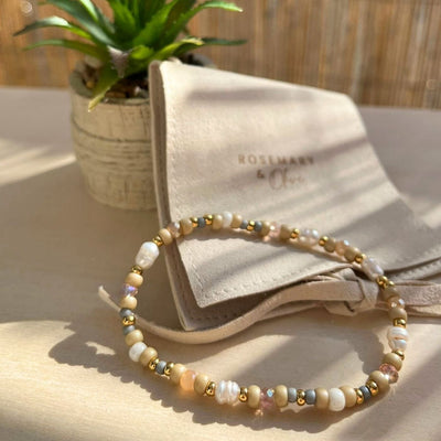 Boho Armband im Sonnenlicht auf heller Holzoberfläche, im Hintergrund ein Schmuckbeutel mit Logo „Rosemary & Olive“ und eine kleine Pflanze.