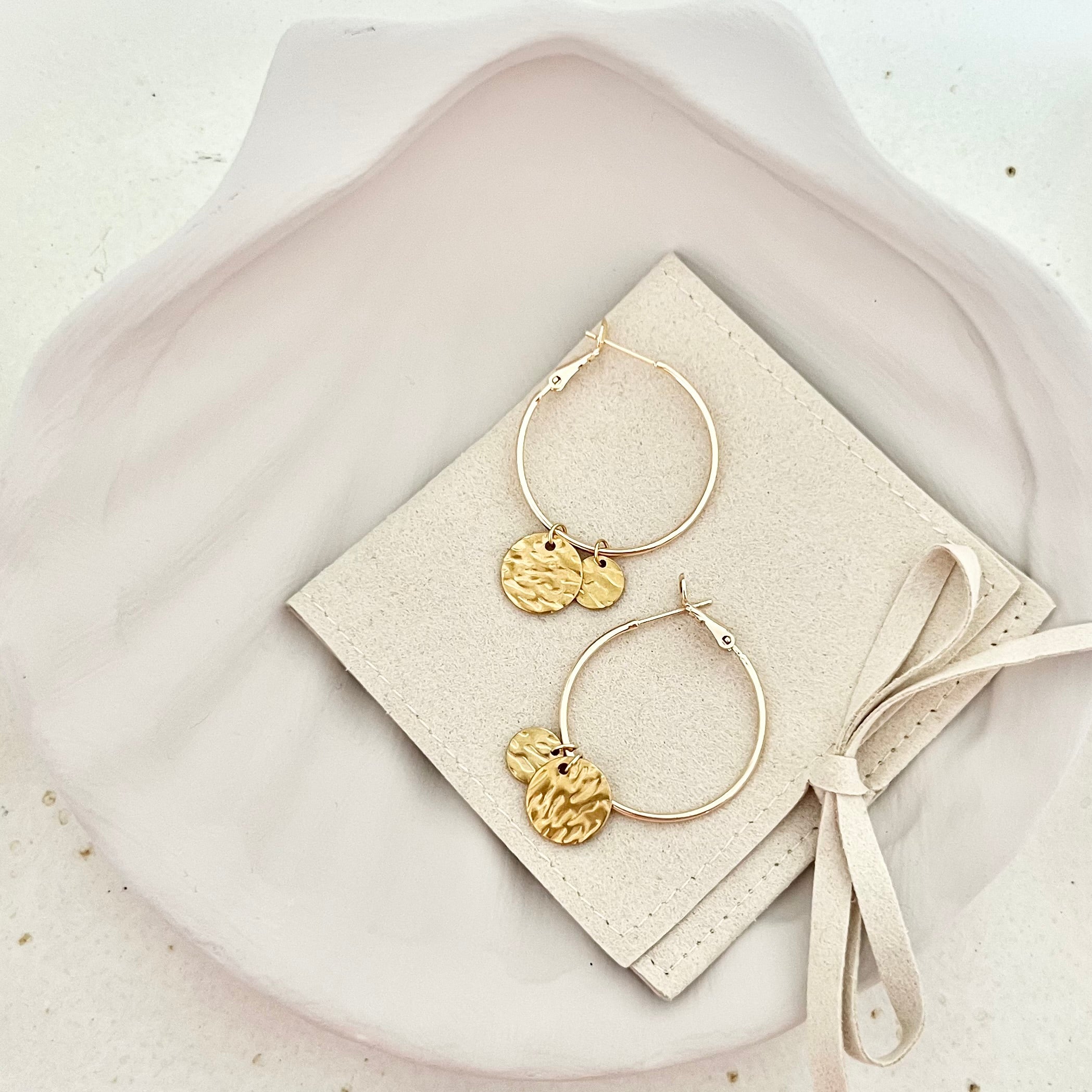 Paar goldfarbene Creolen Ohrringe CASA aus Edelstahl, auf einer Tasche drapiert, das den eleganten und modernen Look des Designs in seiner Gesamtheit zeigt.