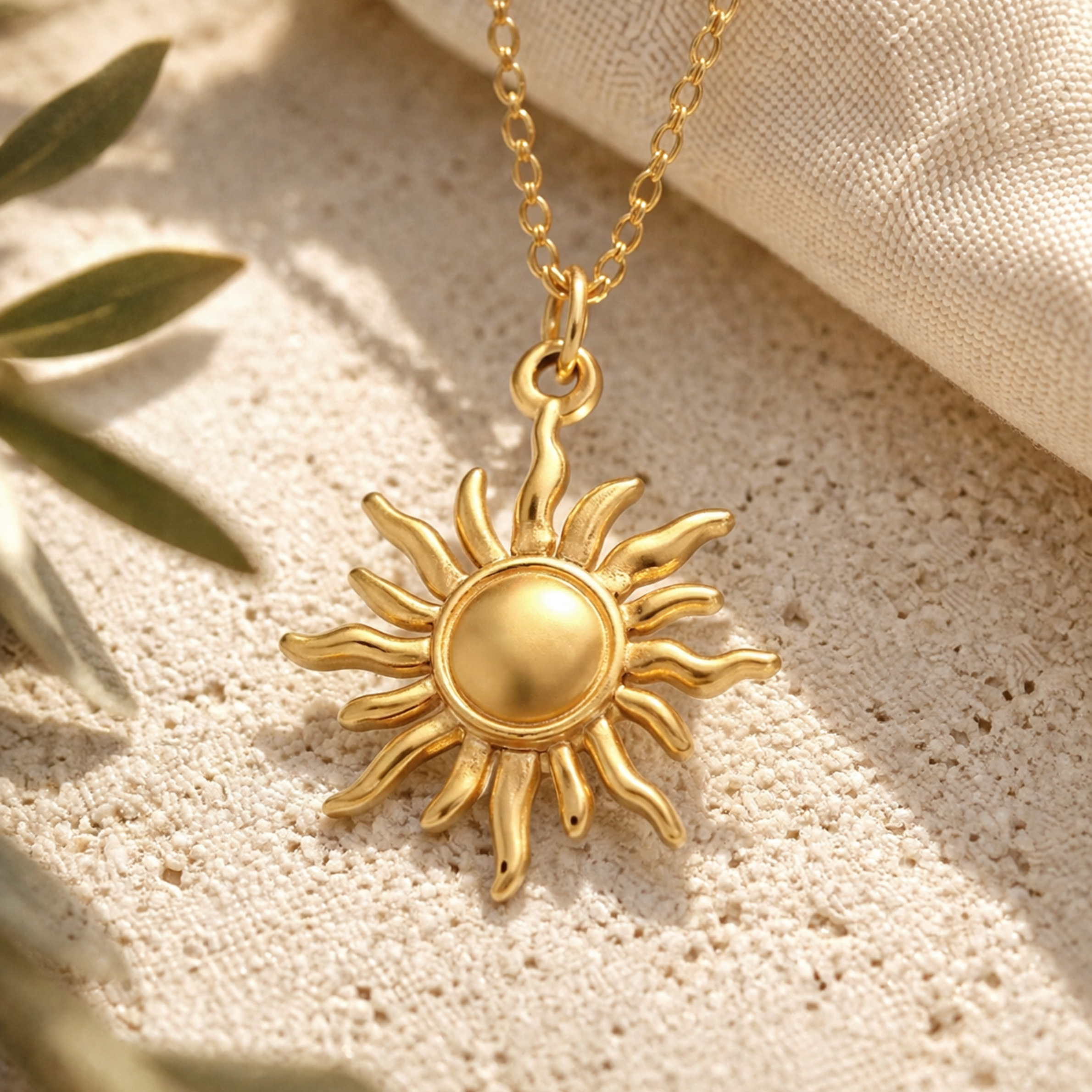 Detailaufnahme einer goldenen Sonnenkette aus Edelstahl auf heller Steinoberfläche mit Leinenstoff und Olivenzweig, Fokus auf glänzenden Anhänger