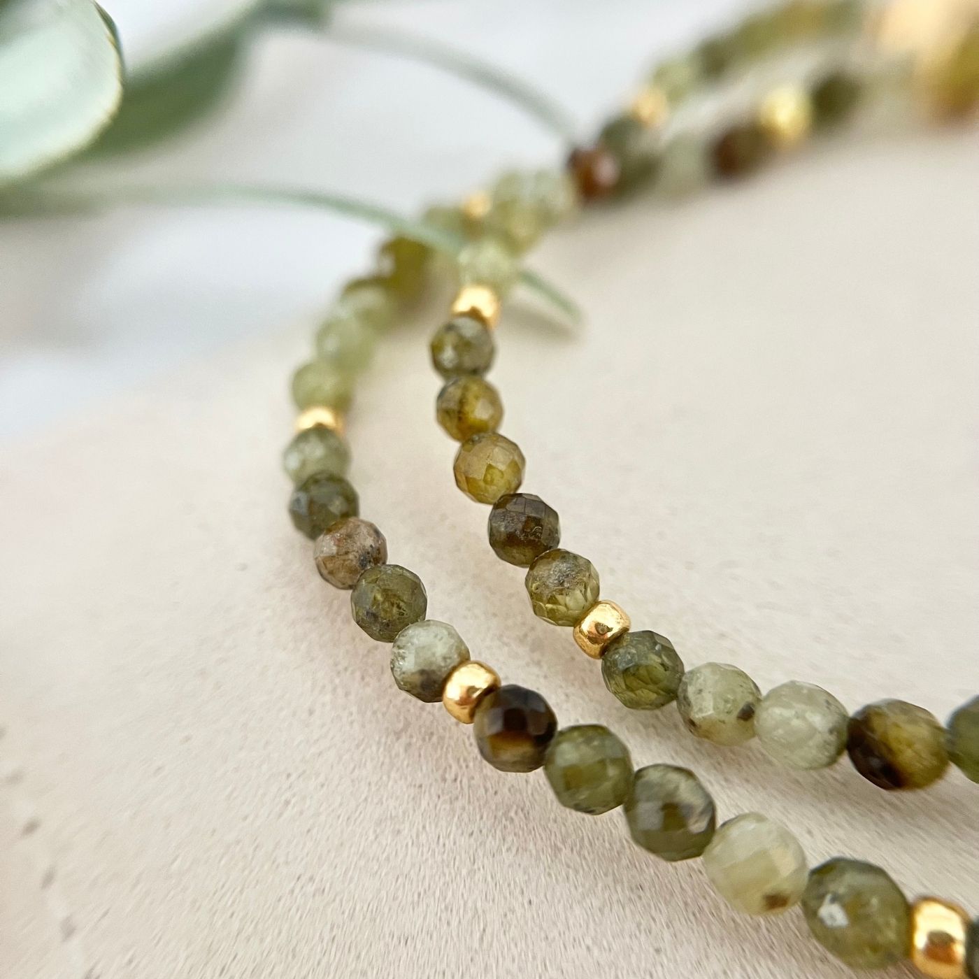 Nahaufnahme zweier Armbänder aus facettierten grünen Natursteinperlen mit goldenen Elementen, auf einem hellen Schmuckbeutel drapiert.