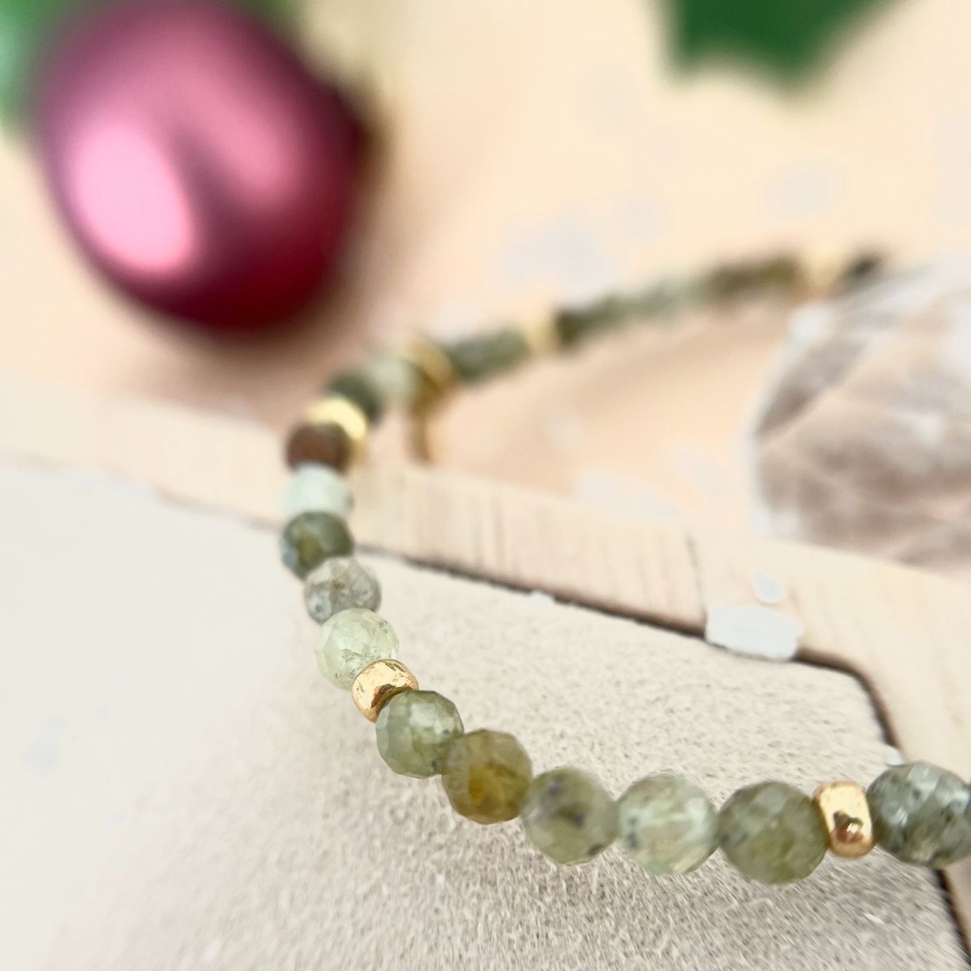 Detailaufnahme eines Armbands mit grünen Natursteinperlen und goldenen Zwischenelementen, im Hintergrund eine rote Weihnachtskugel.
