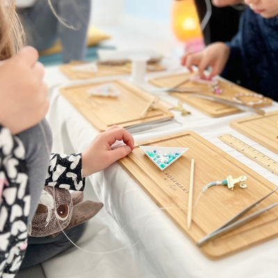Kinderhände beim Fädeln eines Armbands mit bunten Glasperlen auf einem Holztablett – liebevoll gestalteter DIY Schmuck Workshop für Kinder in Wuppertal bei Rosemary & Olive
