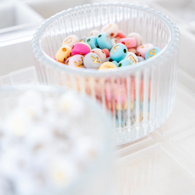 Bunte Buchstabenperlen in Pastellfarben mit goldener Prägung in einer Glasdose – ideal zur Personalisierung von Kinderarmbändern im DIY Schmuck Workshop bei Rosemary & Olive in Wülfrath