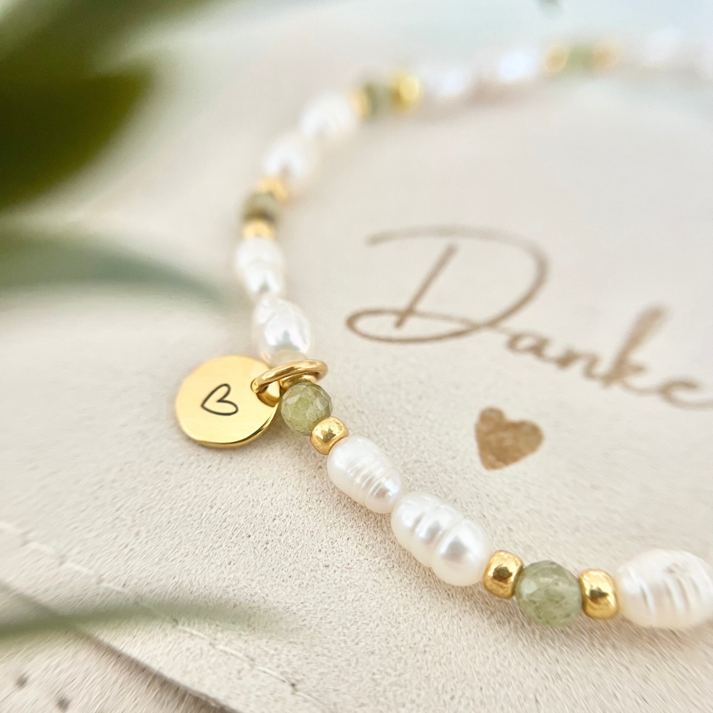 Nahaufnahme eines handgefertigten Perlenarmbands mit weißen Süßwasserperlen, grünen Natursteinperlen und goldenen Details. Der runde vergoldete Anhänger mit Herzgravur liegt auf einer Schmucktasche mit dem Schriftzug „Danke“ – ideales Geschenkarmband für Mama, Freundin oder Trauzeugin.