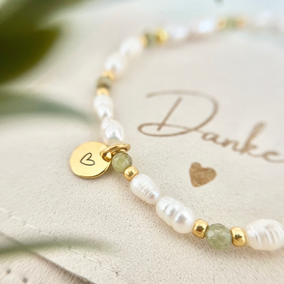 Nahaufnahme eines handgefertigten Perlenarmbands mit weißen Süßwasserperlen, grünen Natursteinperlen und goldenen Details. Der runde vergoldete Anhänger mit Herzgravur liegt auf einer Schmucktasche mit dem Schriftzug „Danke“ – ideales Geschenkarmband für Mama, Freundin oder Trauzeugin.