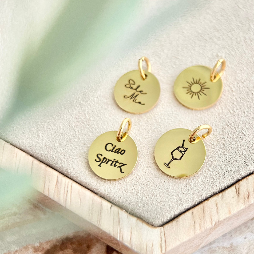 Nahaufnahme vier goldener Anhänger mit Gravuren wie „Sole Mio“, Sonne, „Ciao Spritz“ und Getränkeglas.