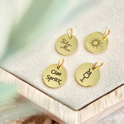 Nahaufnahme vier goldener Anhänger mit Gravuren wie „Sole Mio“, Sonne, „Ciao Spritz“ und Getränkeglas.