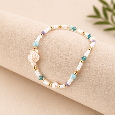 Minimalistisches Produktfoto vom Perlenarmband AMANDA mit weißen, türkisen und goldenen Perlen auf beigem Hintergrund – elastisches Schmuckstück für Frauen
