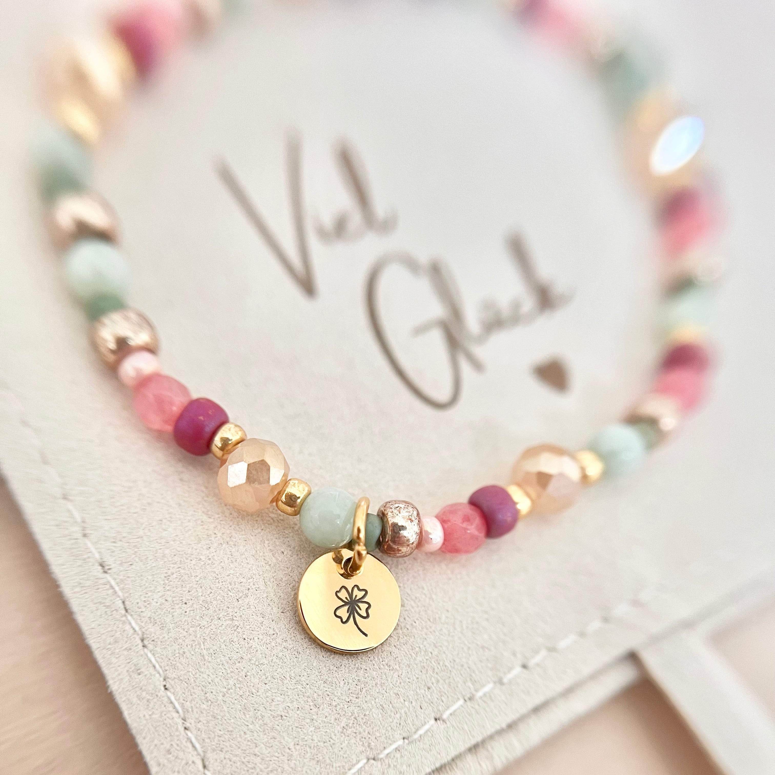 Handgefertigtes Armband FELICIA mit rosa, mintgrünen und goldfarbenen Naturstein- und Glasperlen sowie goldenem Edelstahl-Anhänger mit Kleeblatt-Gravur, präsentiert auf einem Schmuckbeutel mit „Viel Glück“-Schriftzug.