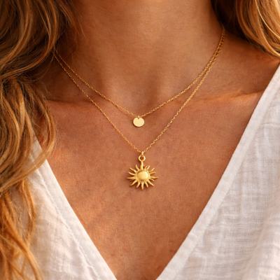 Goldene Sonnenkette aus Edelstahl im Layering Look kombiniert mit feiner goldener Kette mit Plättchen, getragen auf gebräunter Haut mit weißer Bluse