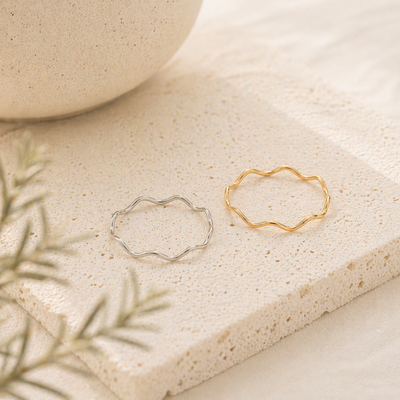 Filigrane Wellenringe in Gold und Silber aus Edelstahl auf heller Steinplatte, minimalistisches Produktfoto mit neutralem Hintergrund, Schmuck für Frauen