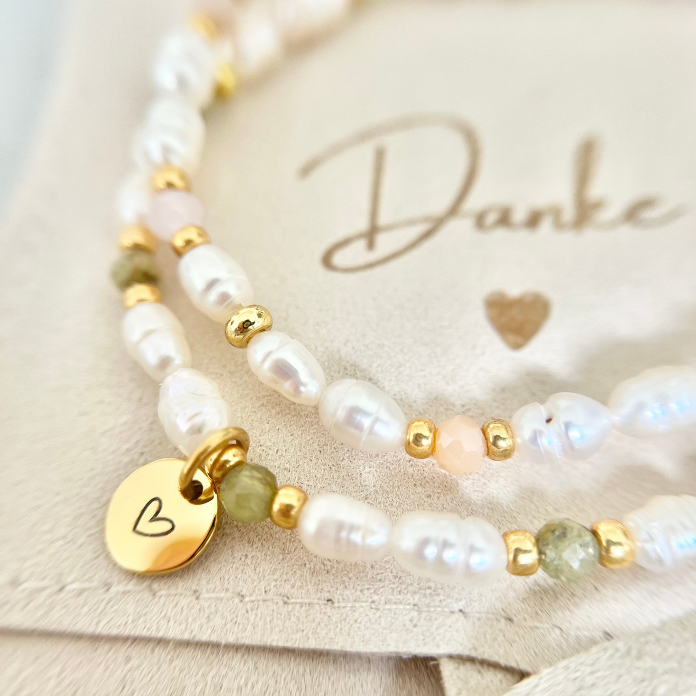 Detailaufnahme eines handgefertigten Perlenarmbands mit weißen Süßwasserperlen, goldfarbenen Zwischenelementen und Glasperlen in Apricot- und Olivgrün. Verziert mit einem runden Edelstahl-Anhänger mit Herzgravur, dekorativ auf Schmuckbeutel mit der Aufschrift „Danke“ – ideales Dankeschön-Geschenk für Trauzeugin, Freundin oder besondere Anlässe.