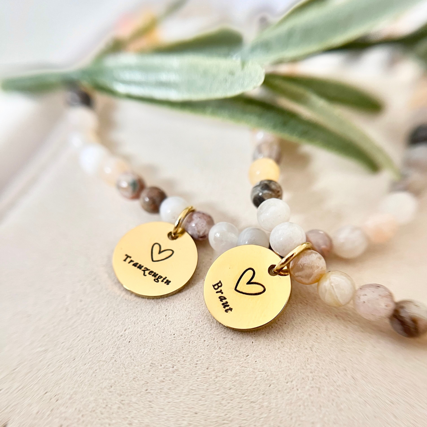 Nahaufnahme von zwei Perlenarmbändern in Weiß-, Beige- und Brauntönen mit runden goldfarbenen Anhängern, graviert mit „Braut“ und „Trauzeugin“, arrangiert als Geschenkset.