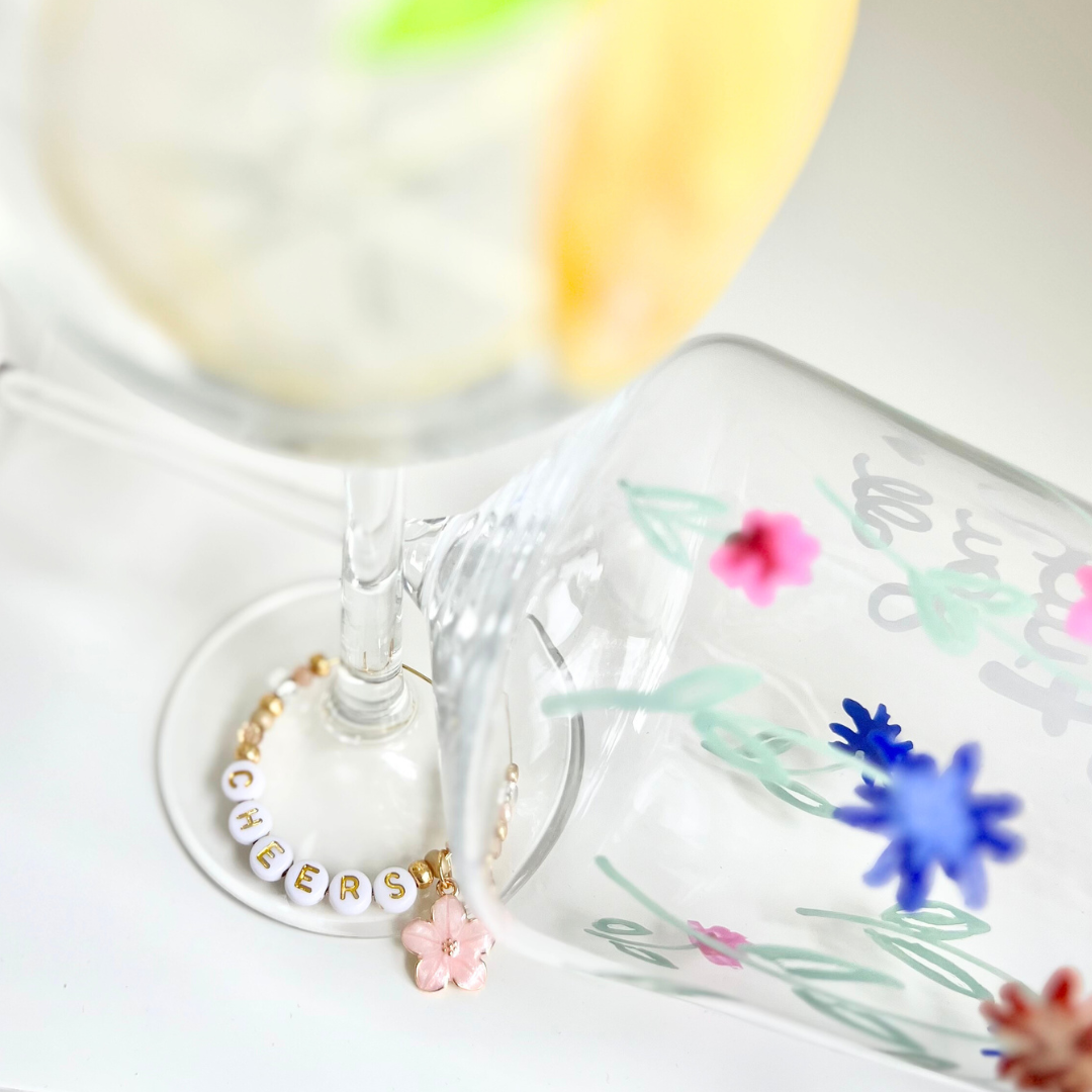 Zwei kreativ bemalte Weingläser: eines mit filigraner und  floraler Malerei in pink, dunkelblau und pastelligem Salbeigrün, das andere mit gemalter Zitrone – ergänzt durch einen personalisierten Glasmarkierer mit ‚CHEERS‘ Buchstabenperlen und rosa Blüten Charm; DIY Projekt aus dem Glass Painting Workshop in Wuppertal und Wülfrath Düssel.