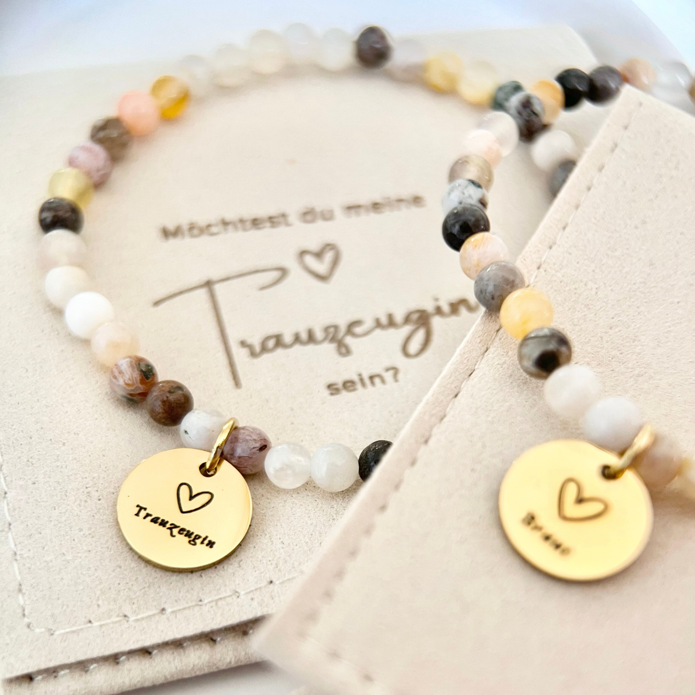 Perlenarmband mit beigefarbenen, weißen und braunen Natursteinen, goldfarbenem Anhänger „Trauzeugin“, platziert auf Schmuckbeutel mit aufgedruckter Frage zur Trauzeugin.