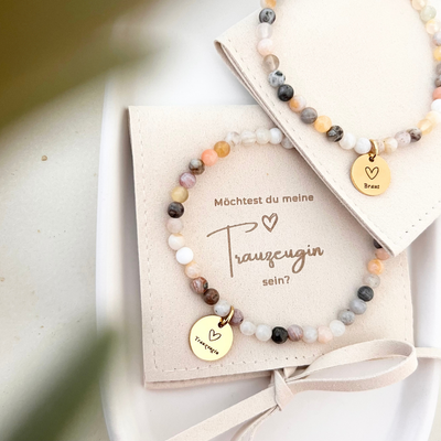 Detailaufnahme eines Perlenarmbands mit weißen und beigen Natursteinen, goldfarbenem Anhänger mit Herzsymbol und Schriftzug „Braut“, dekoriert mit grünem Blatt.