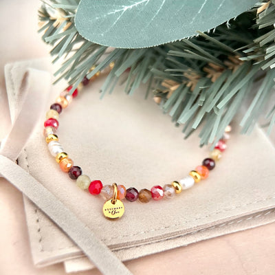 Weihnachtliches Perlenarmband NOELLE von ROSEMARY & Olive mit roten, weinroten, grünen und apricotfarbenen Naturstein- und Glasperlen, weißen Süßwasserperlen und goldenen Akzenten. Das Armband mit goldenem Logoanhänger liegt auf einem cremefarbenen Schmuckbeutel, teilweise bedeckt von Tannenzweigen.
