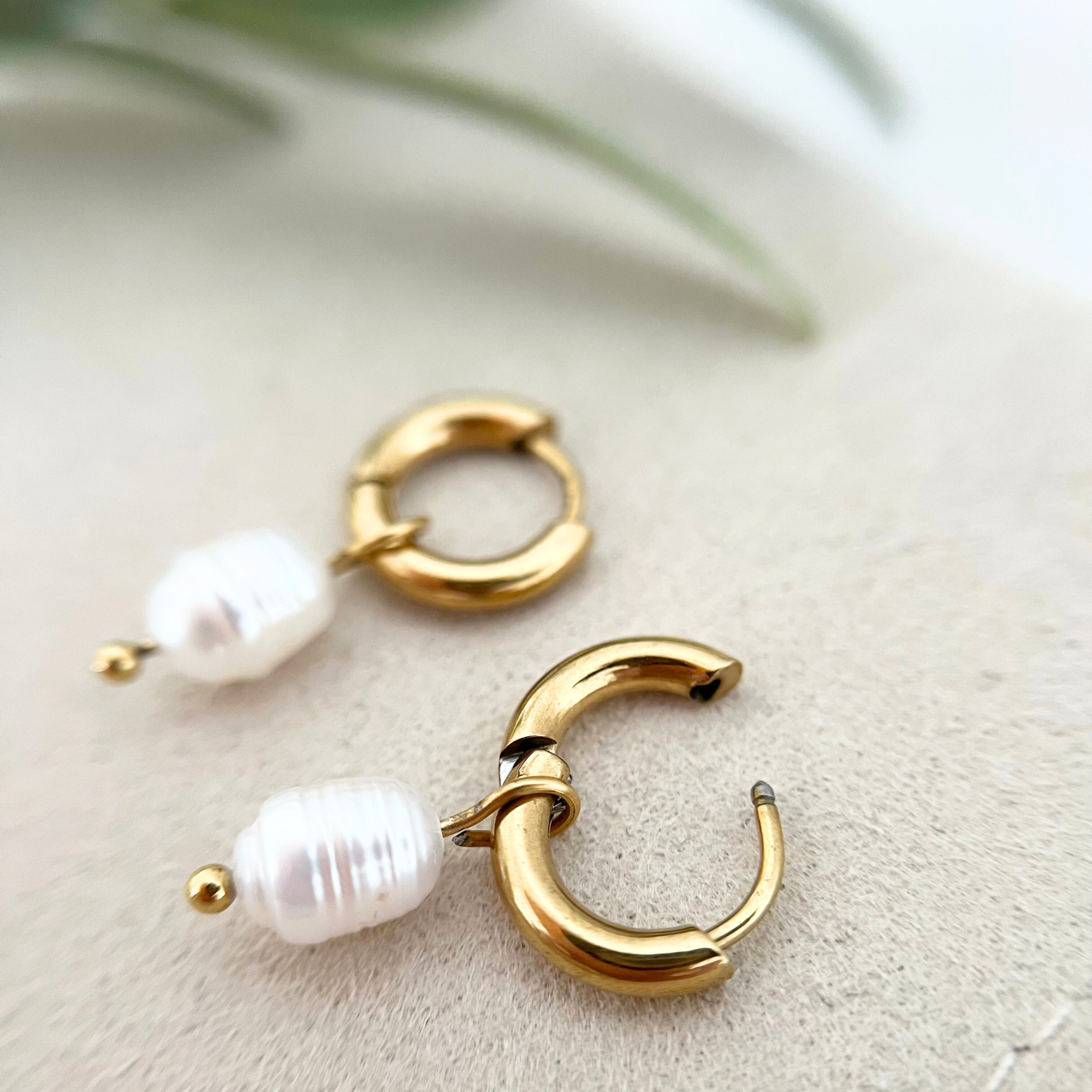 Paar goldene Edelstahl-Ohrringe ISLA mit Süßwasserzuchtperle, auf einer Tasche drapiert, mit einem offenen Ohrring, um den dezenten Charme und das elegante Design zu zeigen.