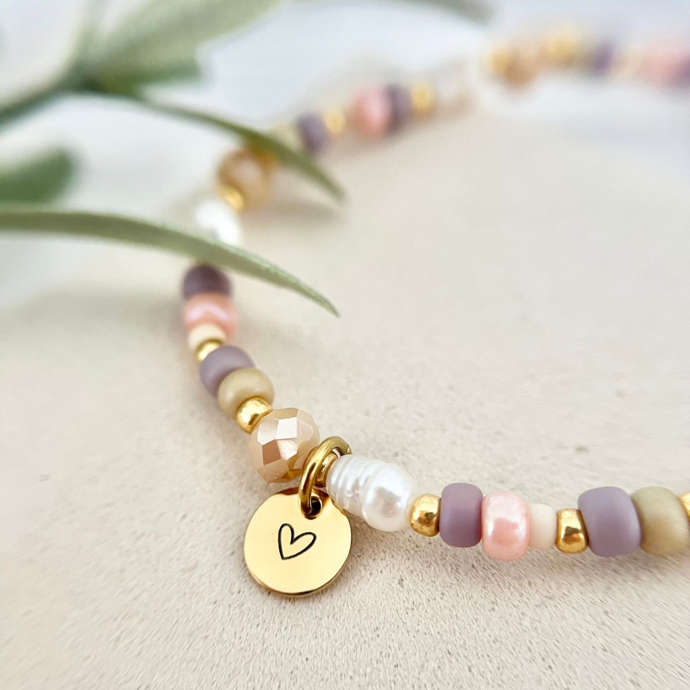 Fröhliches Pastell-Armband mit Herzanhänger aus Edelstahl und Süßwasserperlen, auf Schmuckbeutel drapiert, grünes Blatt im Hintergrund.