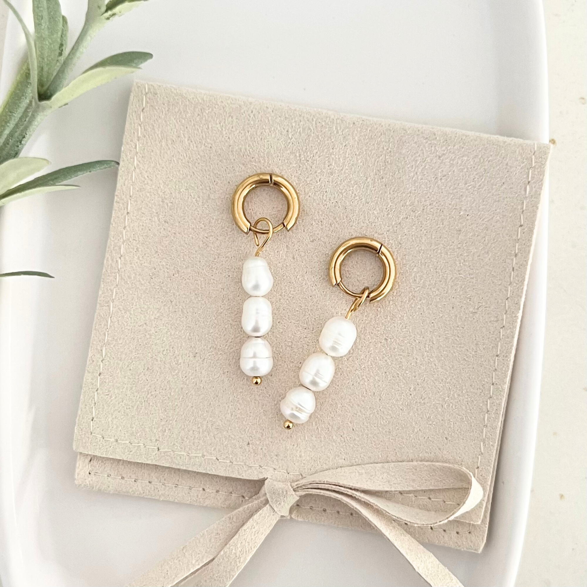 Paar goldene Edelstahl-Ohrringe PERLA mit schimmernden weißen Süßwasserperlen, auf einer Tasche drapiert, um den eleganten Look und die klassische Schönheit hervorzuheben.