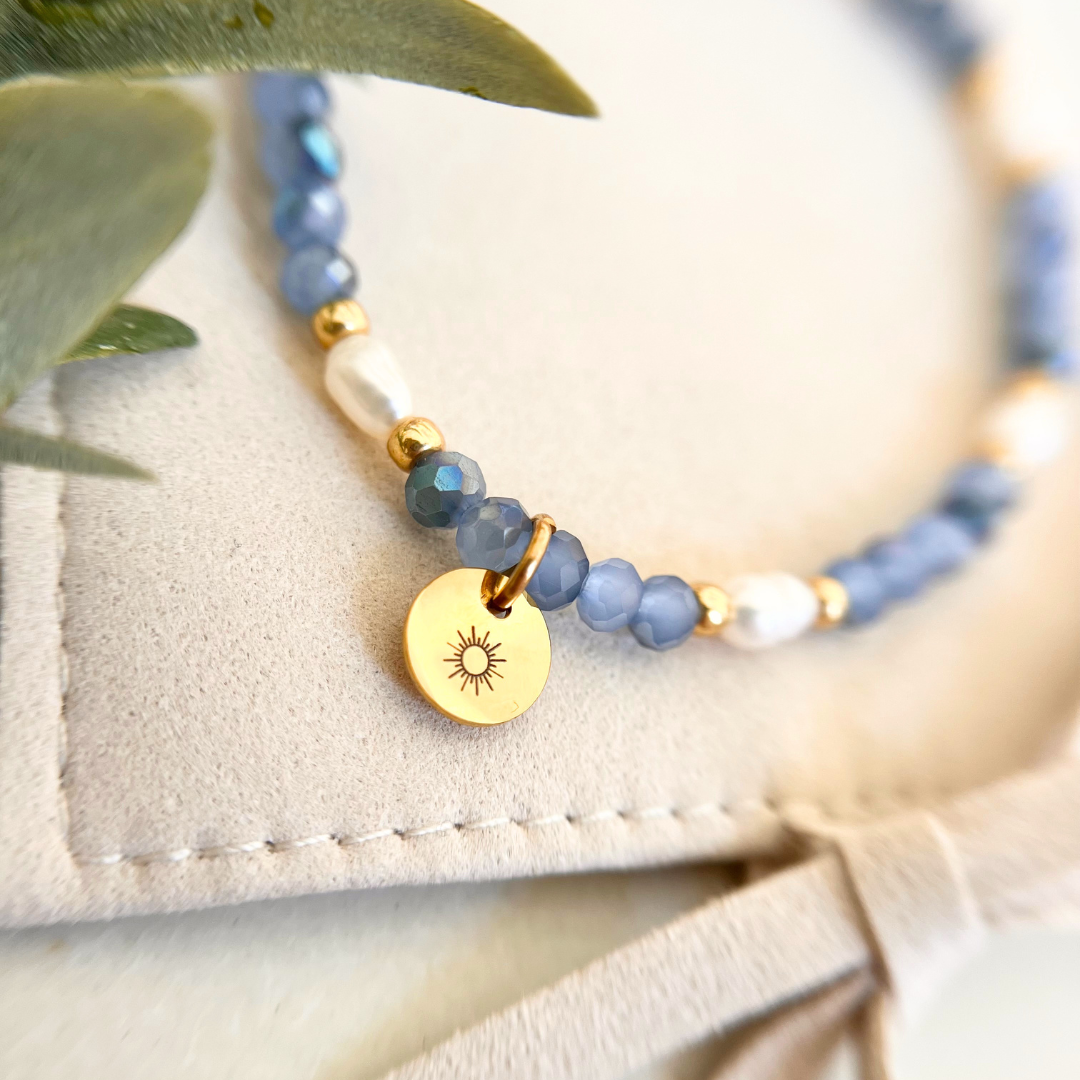 Perlenarmband mit kleinen blauen Facettenperlen, Süßwasserperle und goldenem Sonnenanhänger aus Edelstahl auf hellem Schmuckbeutel, filigranes Schmuckstück für Frauen
