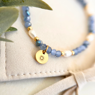 Perlenarmband mit kleinen blauen Facettenperlen, Süßwasserperle und goldenem Sonnenanhänger aus Edelstahl auf hellem Schmuckbeutel, filigranes Schmuckstück für Frauen