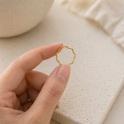 Zarter goldener Wellenring aus Edelstahl wird zwischen zwei Fingern gehalten, natürliche Hautstruktur, heller Hintergrund, minimalistisches Schmuckbild