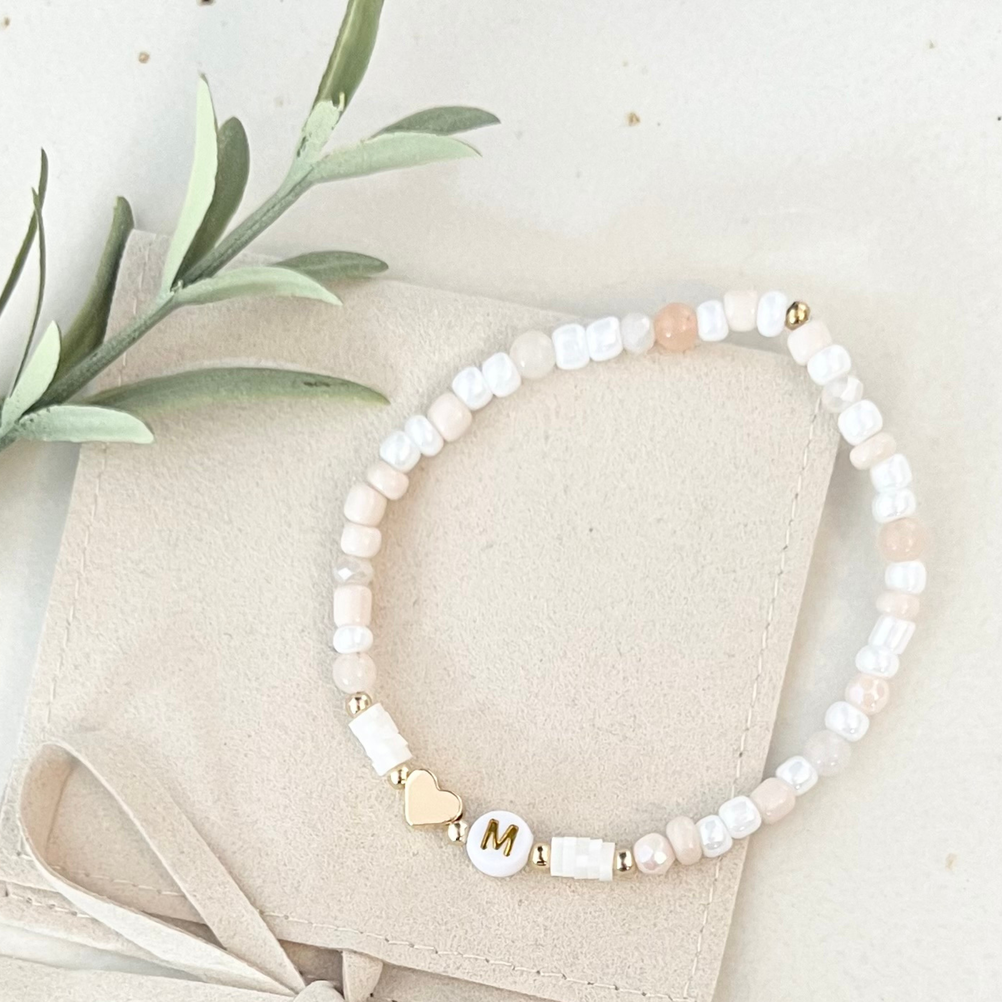 Perlenarmband aus dem DIY Set NATURA mit einem "M"-Anhänger, drapiert auf einer Tasche.